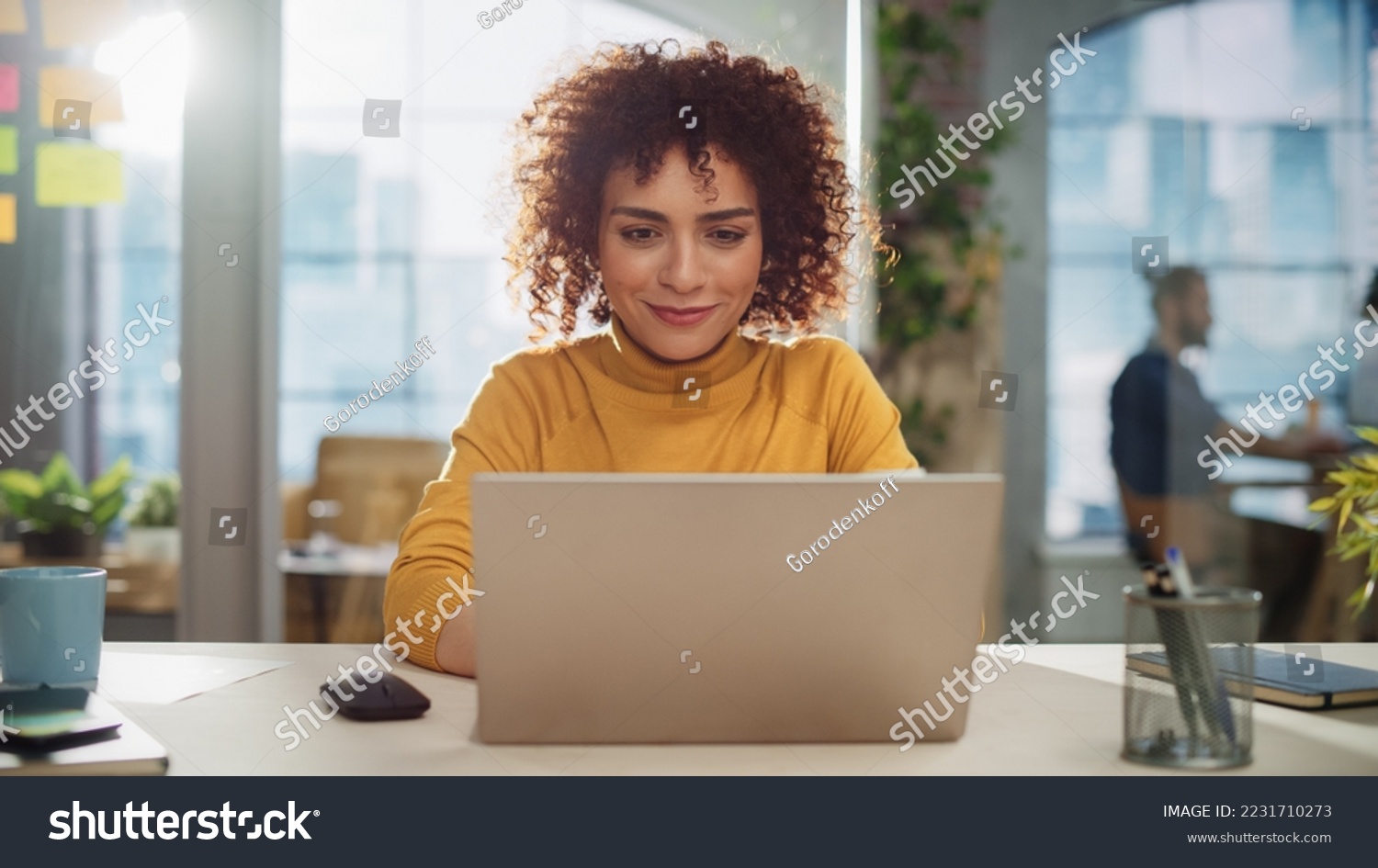 Beautiful Middle Eastern Manager Sitting at a Desk in Creative Office. Young Stylish Female with Curly Hair Using Laptop Computer in Marketing Agency. Colleagues Working in the Background #2231710273