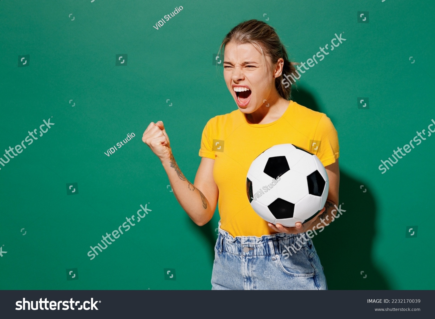 Young angry ardore woman fan wearing basic yellow t-shirt cheer up support football sport team hold in hand soccer ball watch tv live stream scream clench fist isolated on dark green background studio #2232170039