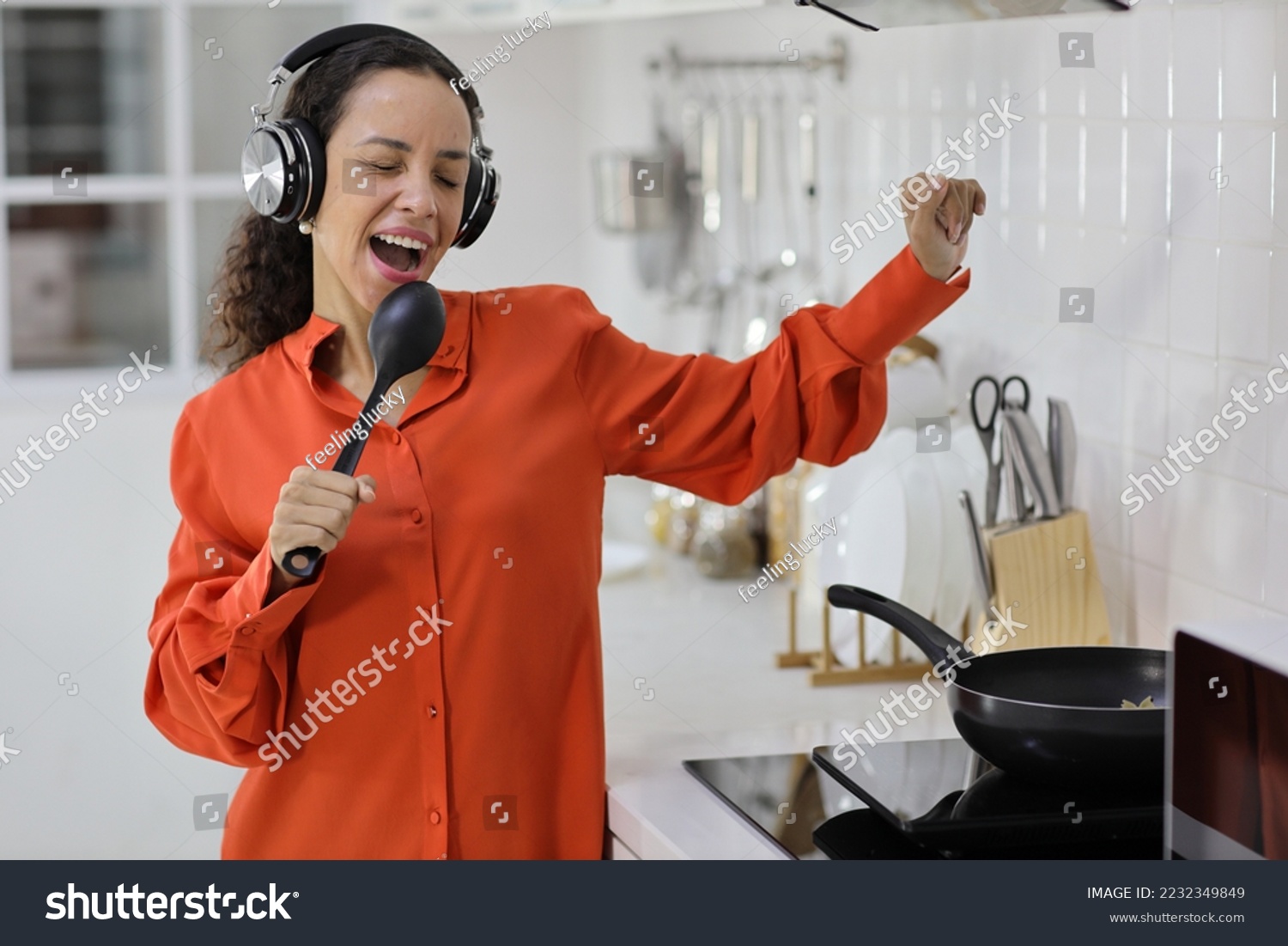 Happy latin woman cooking, smelling and tasting food while dancing in kitchen. Beautiful young female with headphone preparing delicious meal while listen music at home. Healthy and lifestyle concept #2232349849