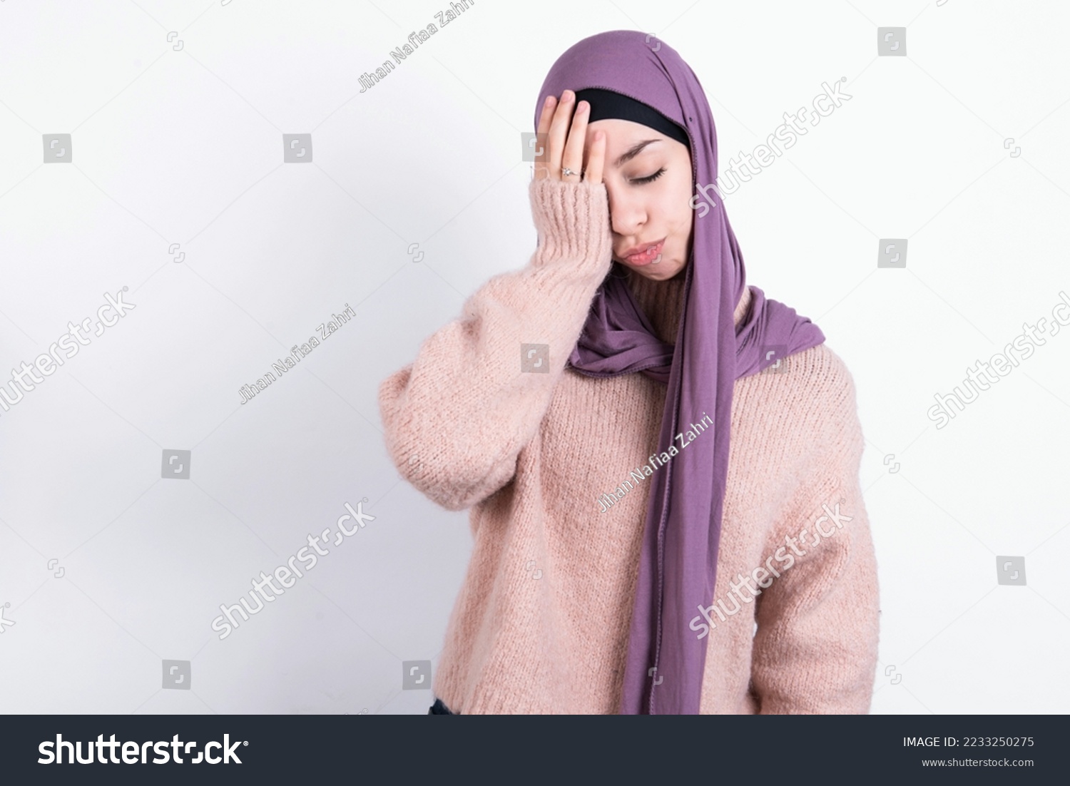 Tired overworked muslim woman wearing hijab and knitted sweater over white background has sleepy expression, gloomy look, covers face with hand, has eyes shut, gasps from tiredness, fatigue  #2233250275