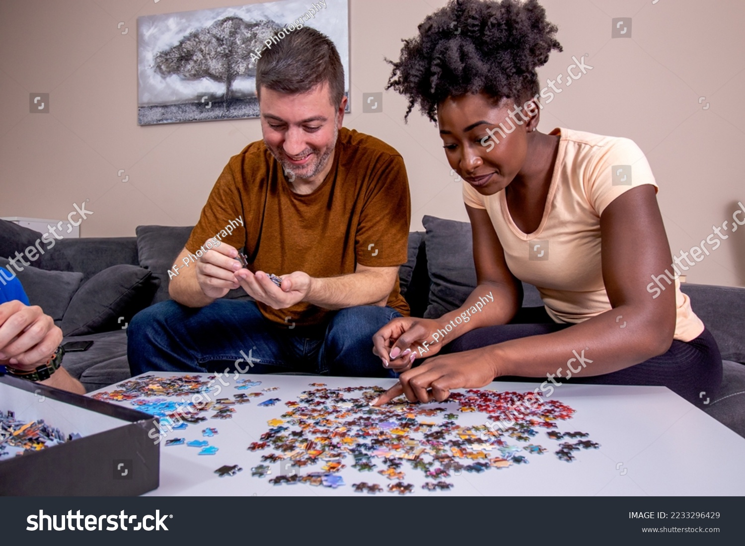 A couple playing with a jigsaw puzzle at home, on a white wooden table. Putting things together and solving problems. Fun and diversity in friendship. #2233296429