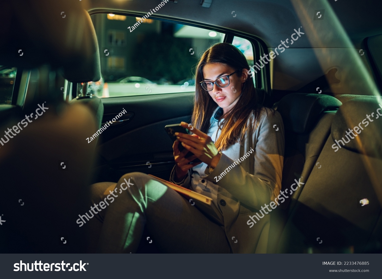 Beautiful young businesswoman traveling with car on a business trip during night while sitting in a back seat and using a smartphone. Gorgeous female using mobile phone to send email or messages. #2233476885