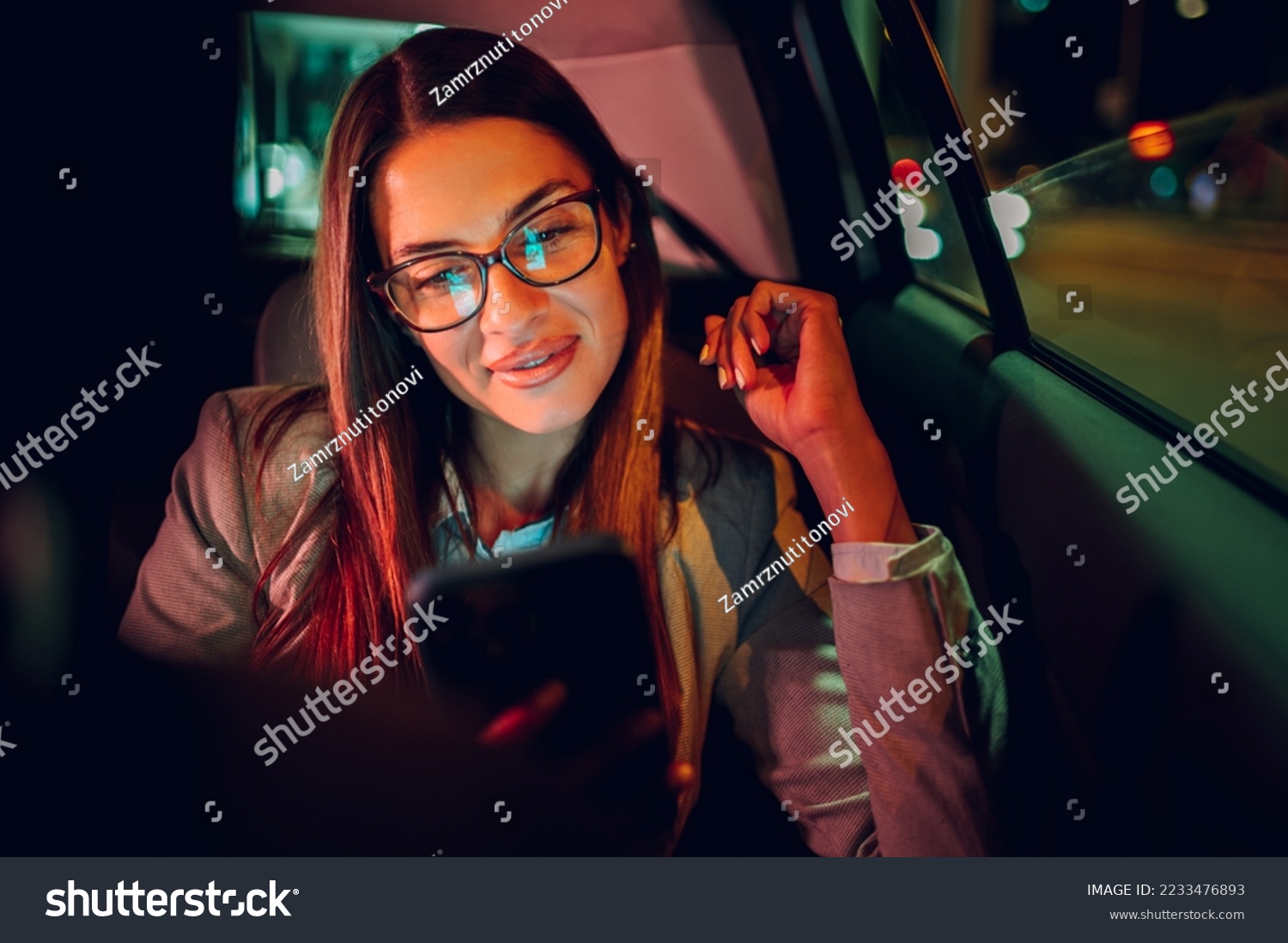 Portrait of a businesswoman commuting from office in a taxi backseat and using a smartphone in city at night after late work. Beautiful female using mobile phone while sitting on a backseat of a car. #2233476893