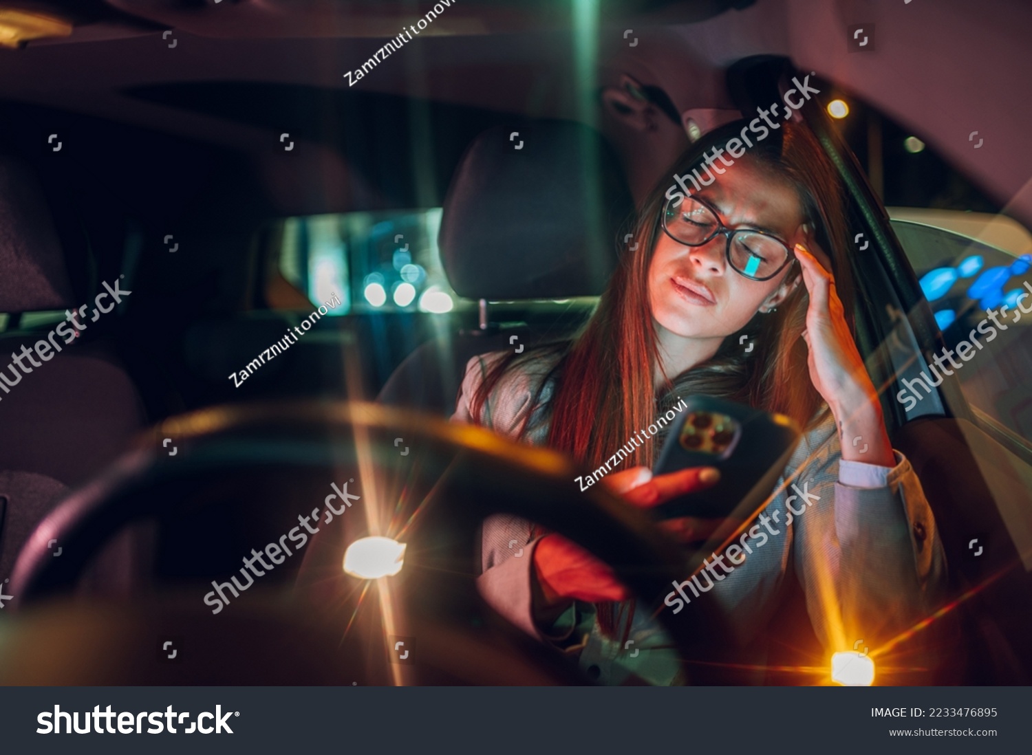Businesswoman driver sitting in car checking mobile phone while riding in urban city. Young woman looking at smartphone while commuting from work at night. Having a headache and feeling tired. #2233476895