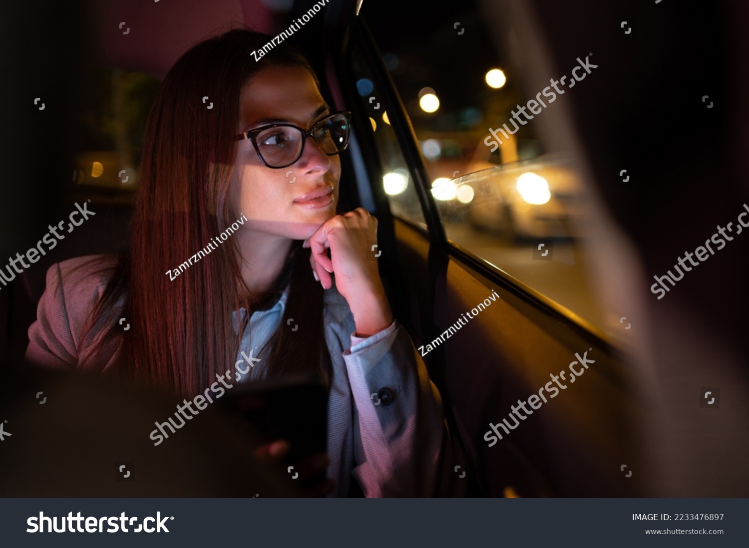 Successful young businesswoman in fashion office clothes working late and using smartphone and looking trough the window while sitting in a backseat of car in urban modern city during night. #2233476897