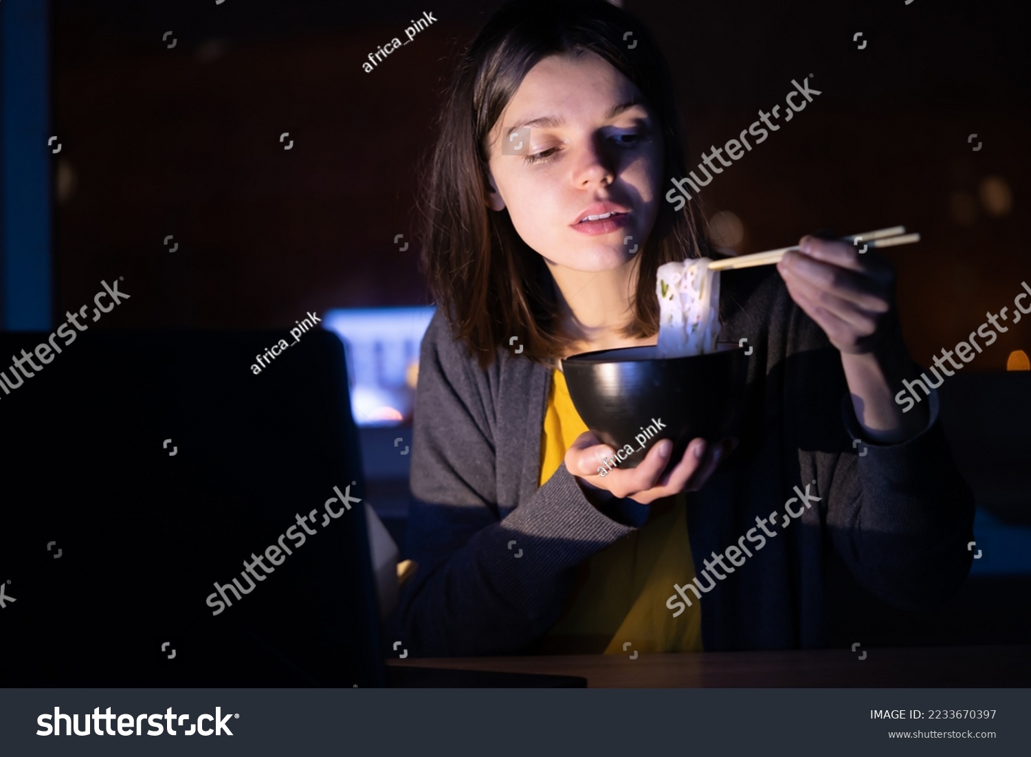 Young girl works at night at home with laptop and eats noodles, asian food with chopsticks. A woman watches a show, a movie and has dinner with ordered food from a cafe. #2233670397