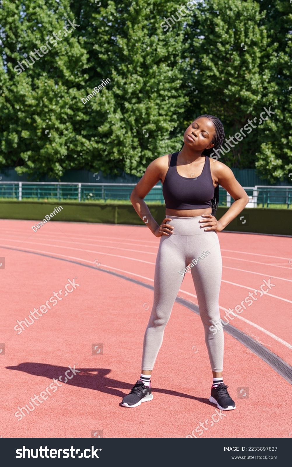 African American woman wearing grey leggings and black top prepares for competition. Black sportswoman stretches body with hands on hips for running marathon on red jogging track in sunny morning #2233897827