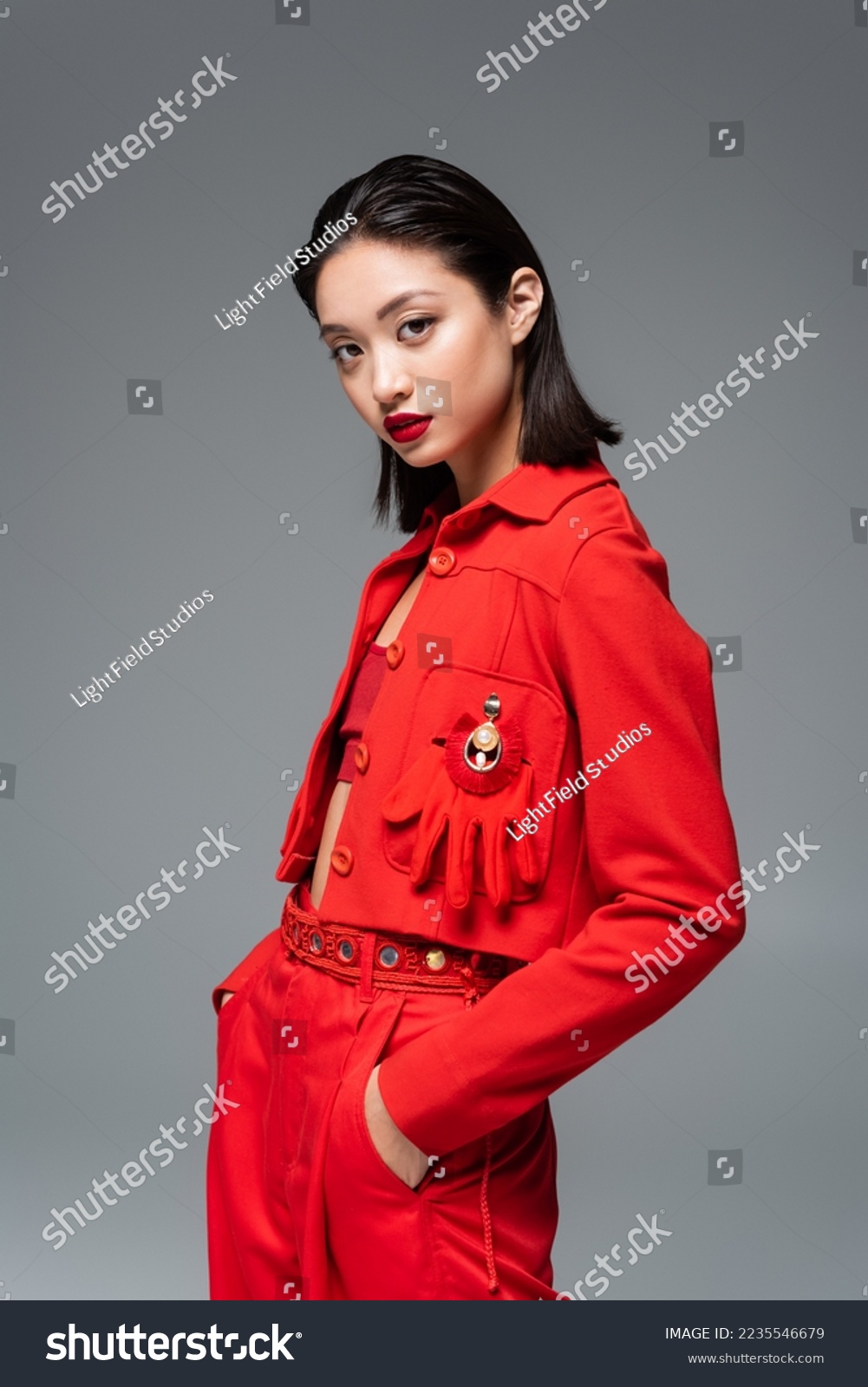 asian woman in red jacket decorated with brooch and glove standing with hands in pockets isolated on grey #2235546679