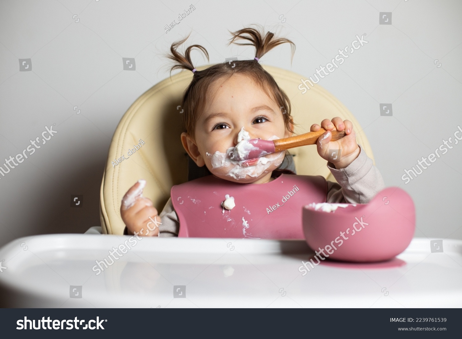 Small cute little toddler brunette caucasian girl with two tails tasting and enjoying by herself with a spoon the greek yogurt sitting in baby chair with messy face; self-feeding concept #2239761539