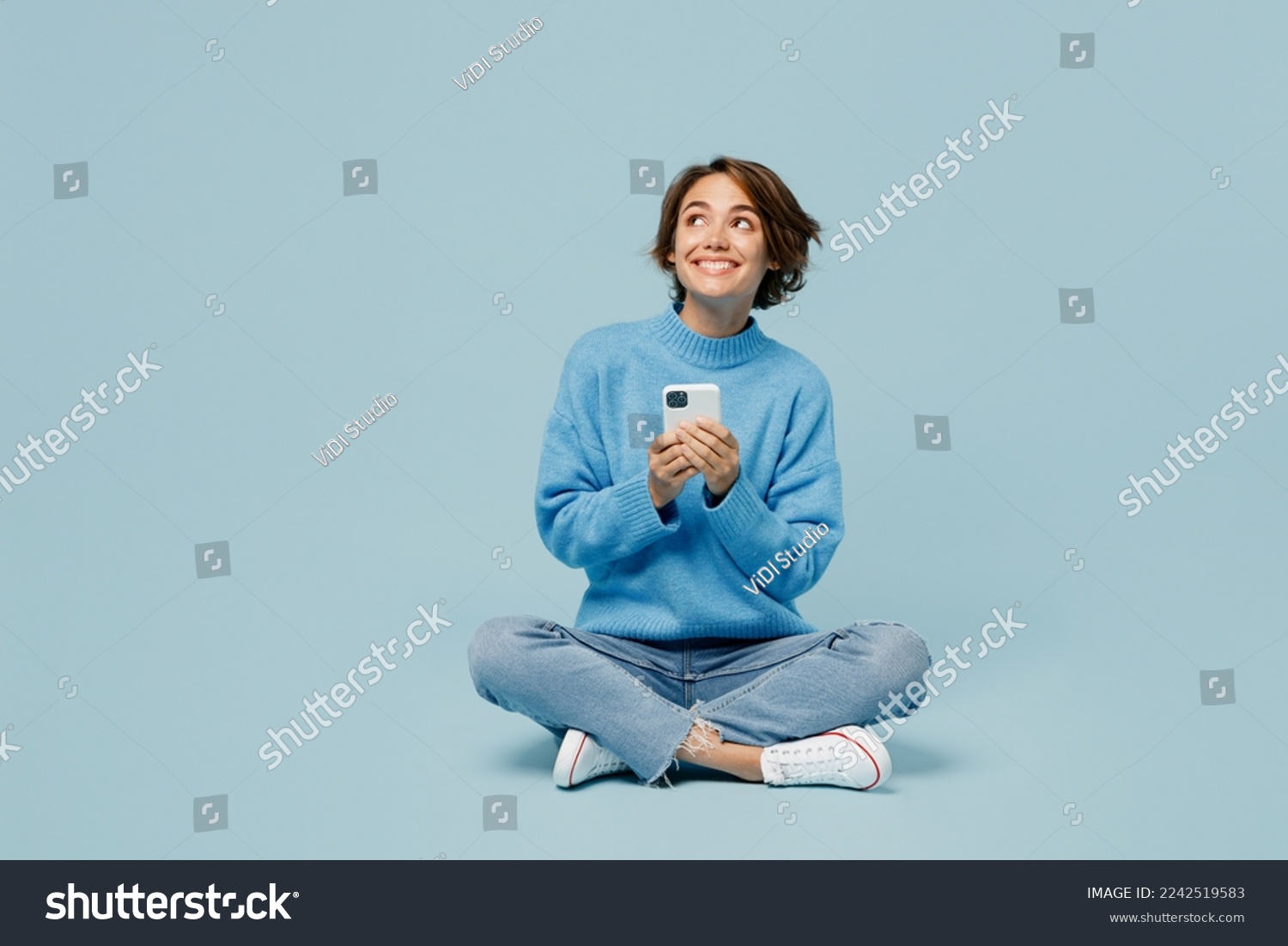 Full body fun young woman wear knitted sweater hold in hand use mobile cell phone look aside on workspace isolated on plain pastel light blue cyan background studio portrait. People lifestyle concept #2242519583