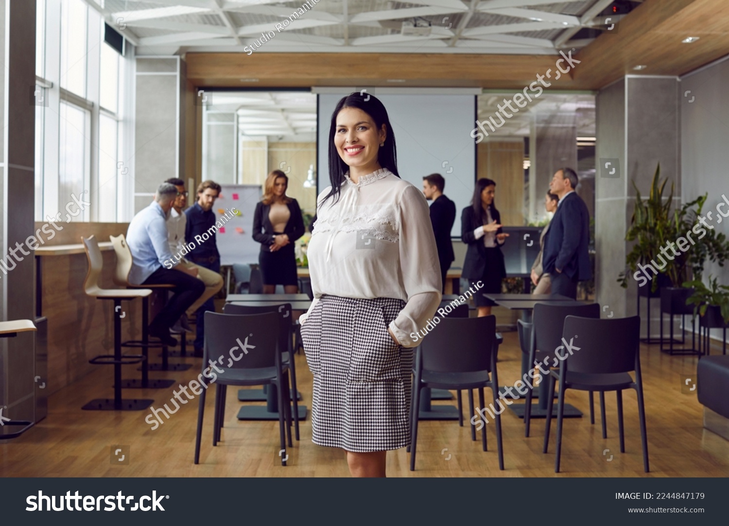 Portrait of friendly and successful business woman standing against background of colleagues in office. Millennial female leader or coach stands against background of people talking to each other. #2244847179