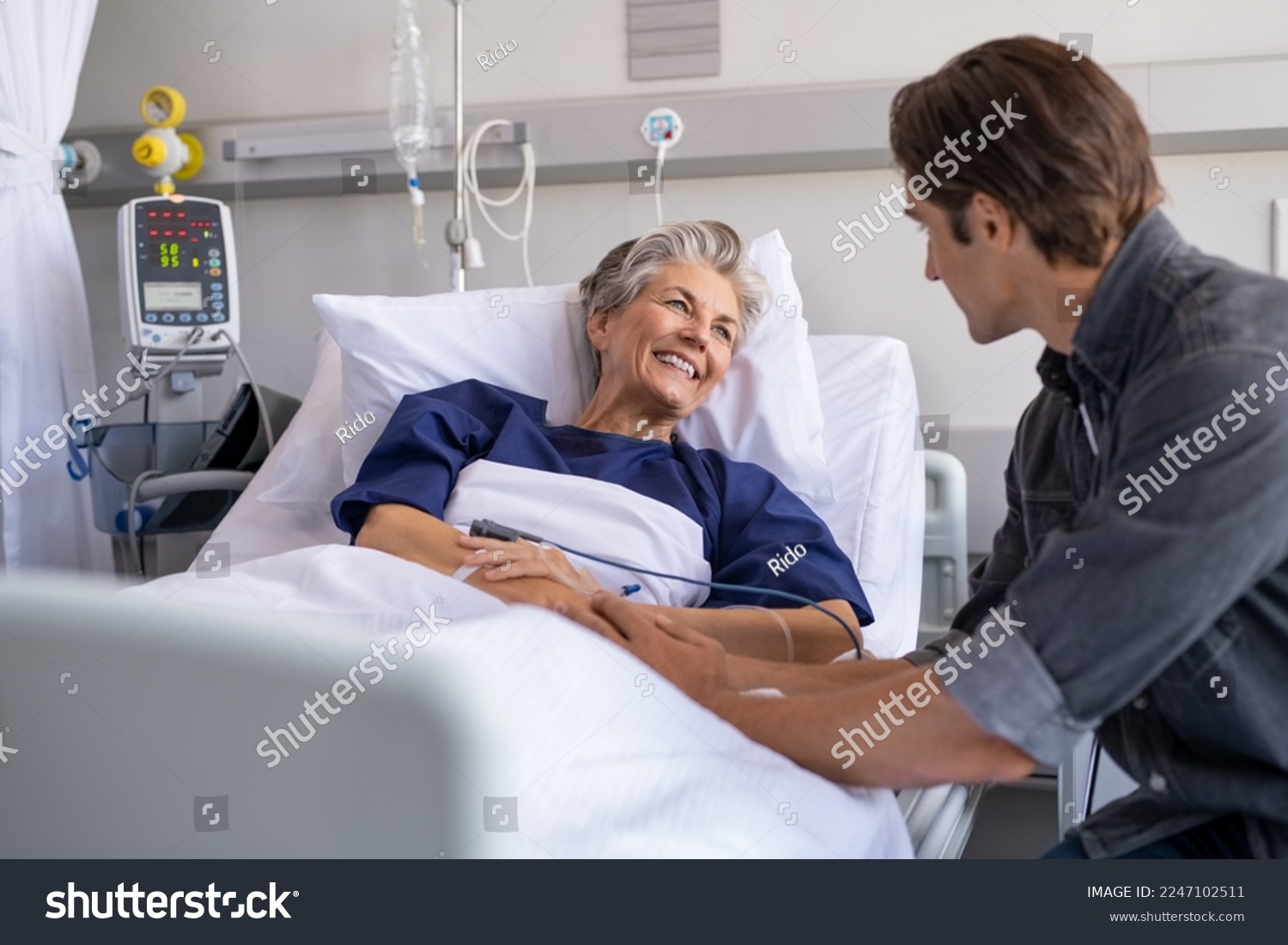 Son visiting sick old mother in hospital. Smiling senior patient in conversation with man. Grandson visiting and cheering his happy granny lying in bed at modern hospital ward. #2247102511
