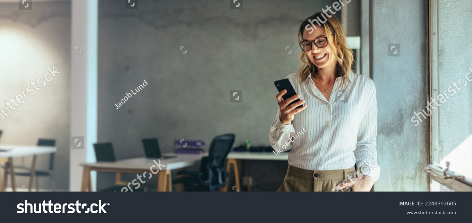 Smiling businesswoman using her phone in the office. Small business entrepreneur looking at her mobile phone and smiling while communicating with her office colleagues #2248392605