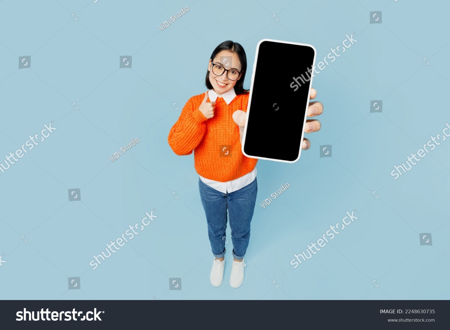 Full body top view from above young woman of Asian ethnicity wear orange sweater glasses hold in hand use mobile cell phone with blank screen area isolated on plain pastel light blue cyan background #2248630735