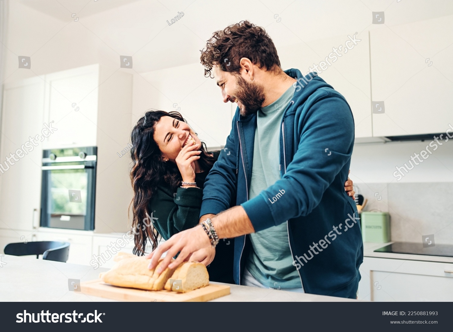 A young caucasian couple in their 30s shares a laugh while they eat bread in a modern, bright kitchen. He's cutting bread, she's smiling and biting a slice of bread. Domesticity and joy lifestyle #2250881993