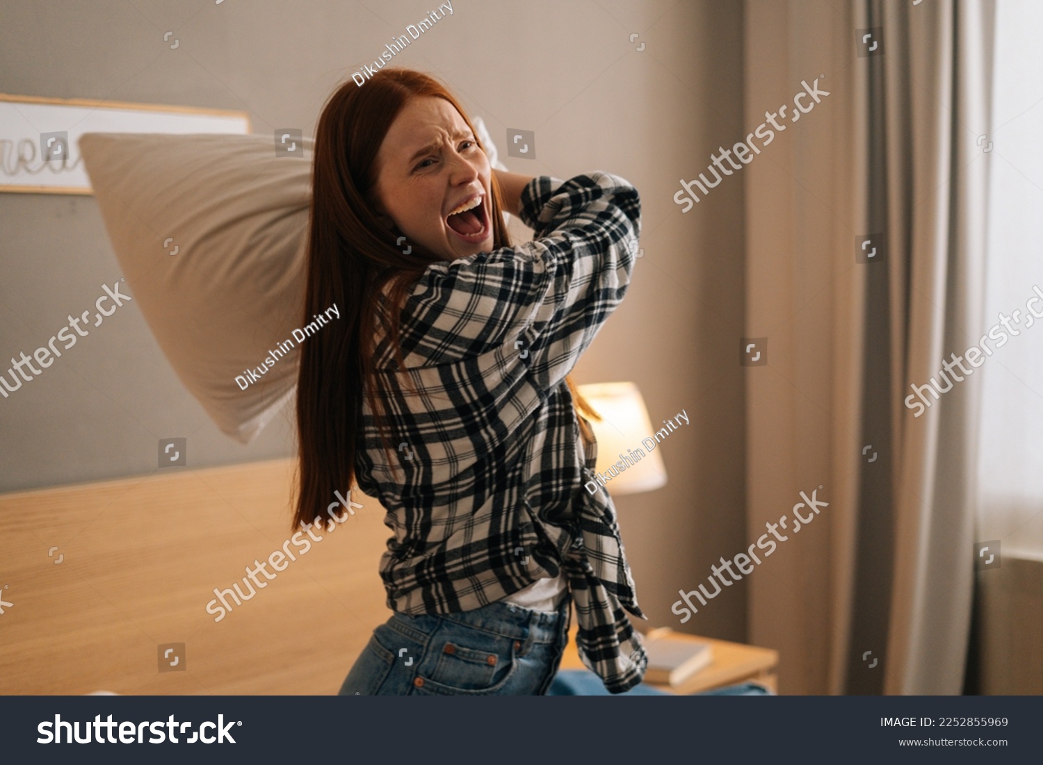Portrait of angry annoyed woman with open mouth shouting and yelling with aggressive expression, throwing pillow sitting on bed in bedroom. Furious crazy redhead female vented anger in privacy of home #2252855969