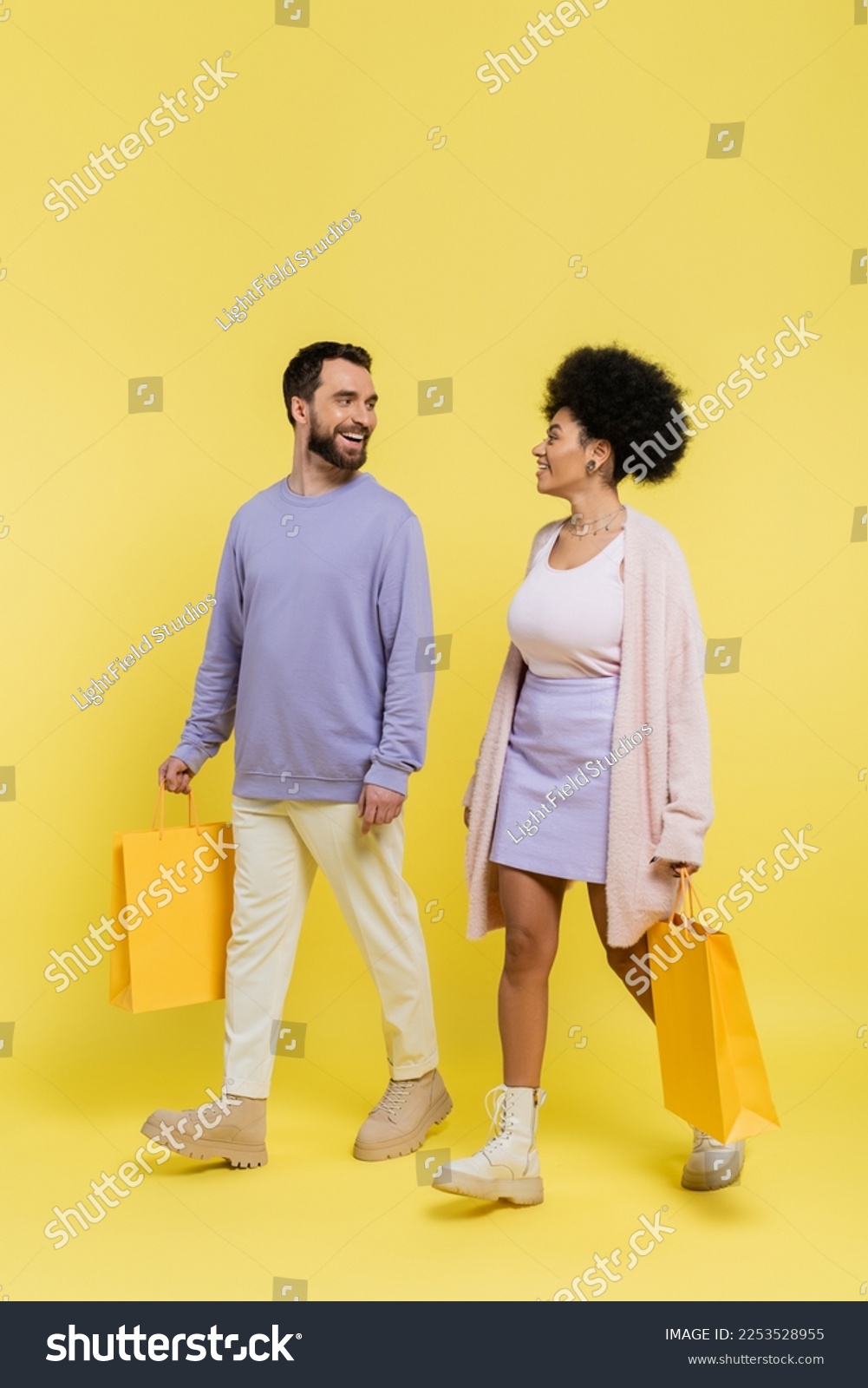 full length of happy and stylish interracial couple walking with shopping bags and looking at each other on yellow background #2253528955