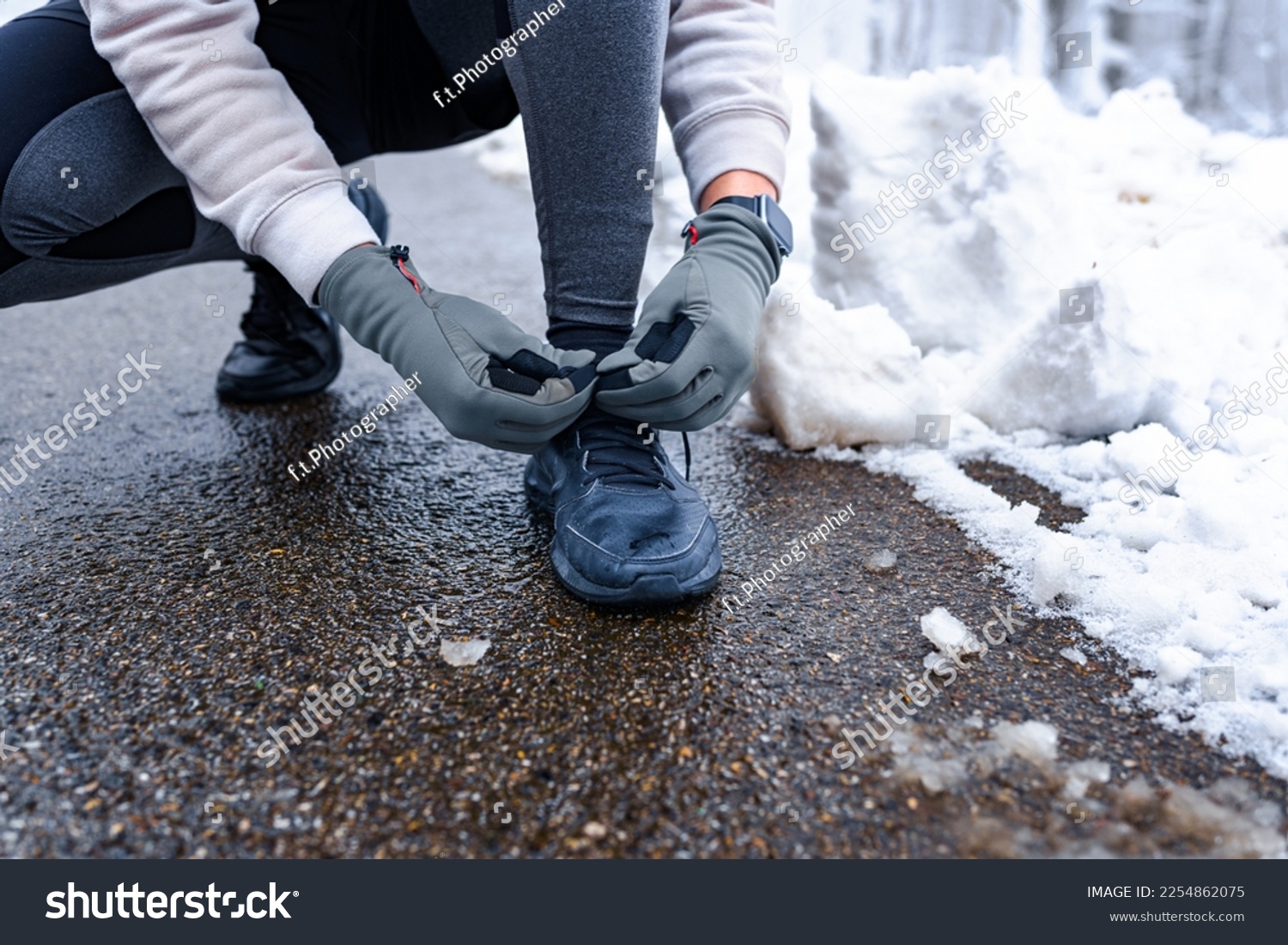 Closeup image of a person tying shoelaces on wet road, snow on the ground and sports equipment in focus. #2254862075