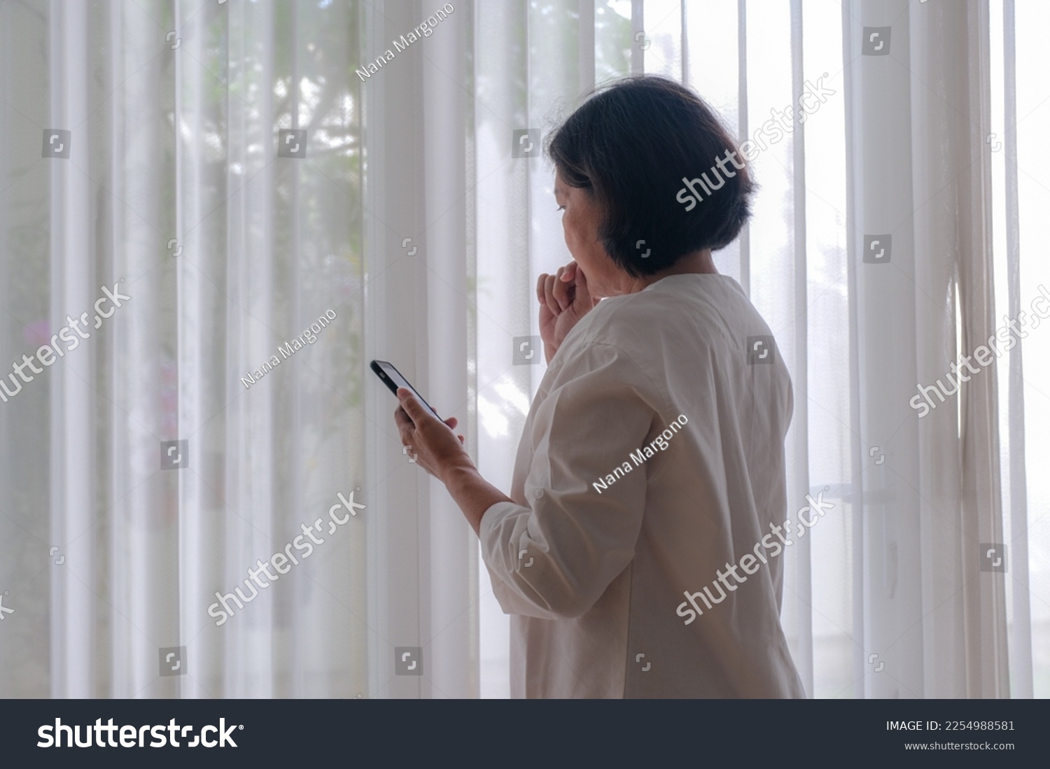 Back view: A woman holding a cell phone standing near the window; biting nails. #2254988581