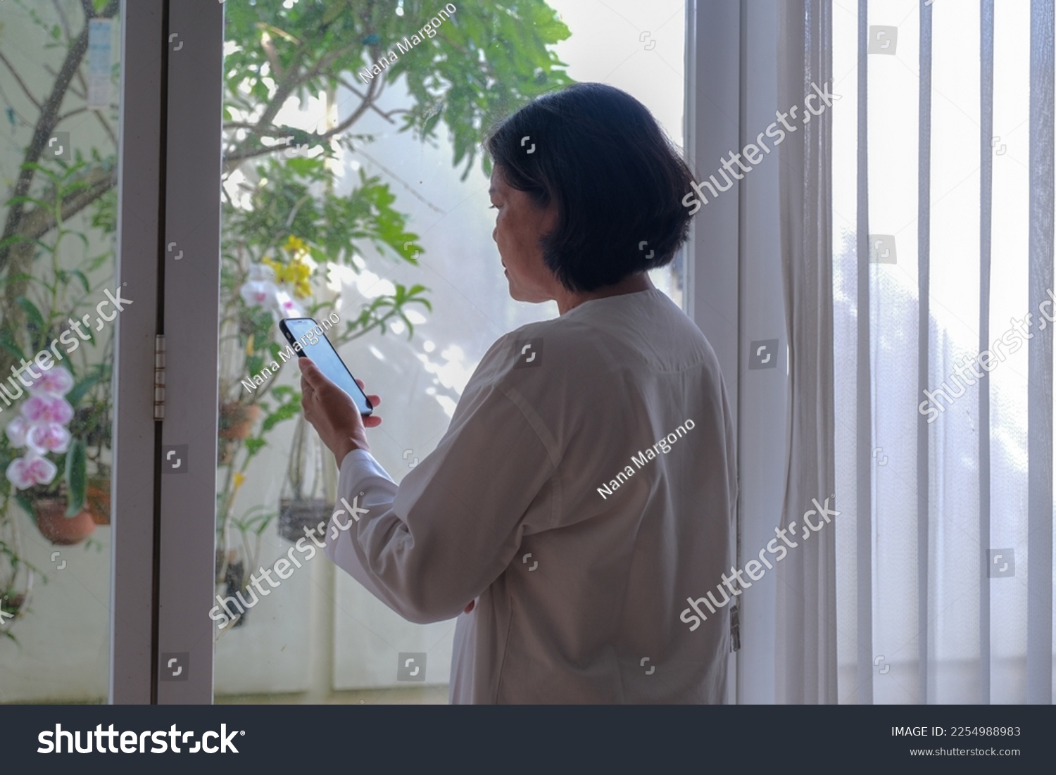 Back view: A woman holding a cell phone standing near the window #2254988983