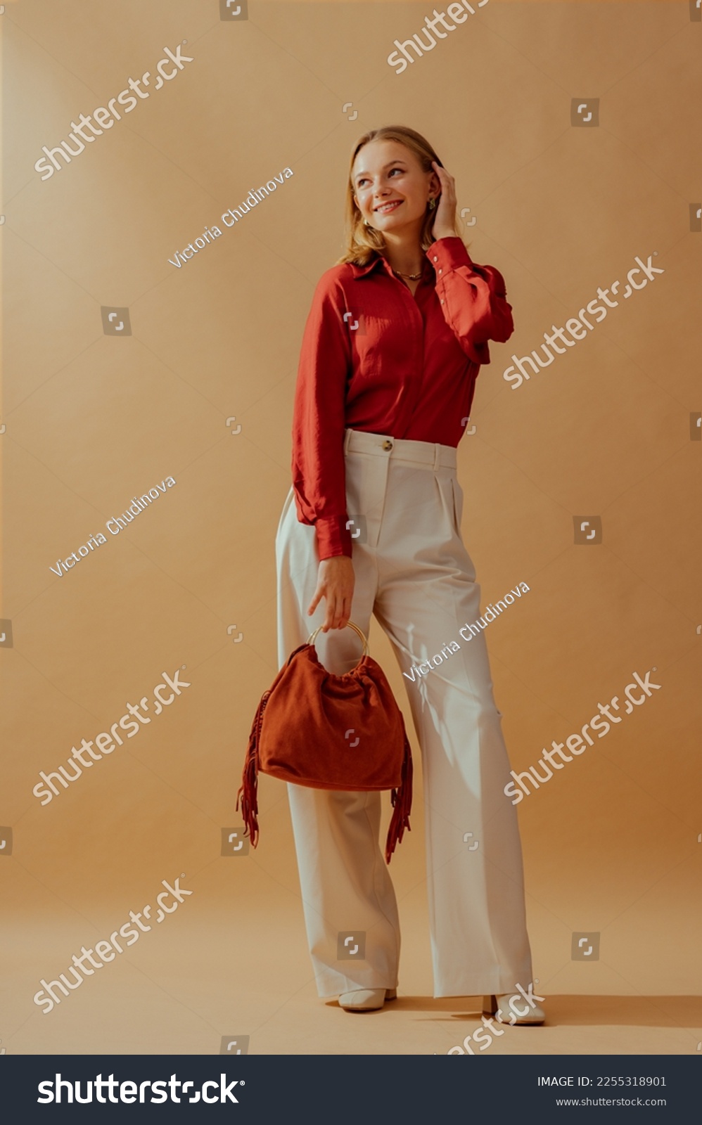 Happy smiling fashionable woman wearing elegant satin blouse, white wide leg trousers, holding trendy suede fringed bag, posing on beige background. Full-length studio fashion portrait #2255318901