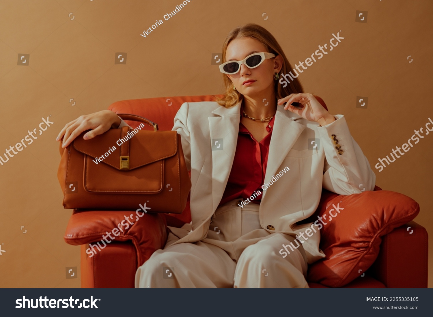Fashionable confident woman wearing elegant white suit, sunglasses, chunky chain, holding classic brown leather bag, sitting in armchair, posing on beige background. Copy, empty space for text #2255335105