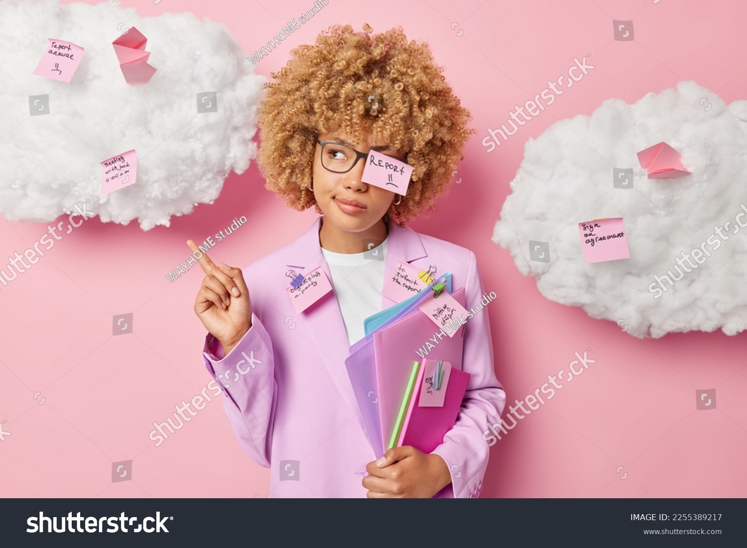 Serious female office worker coveres with memo stickers writes down list to do poses with folders indicates index finger overhead dressed in formal clothes isolated over pink background clouds above #2255389217