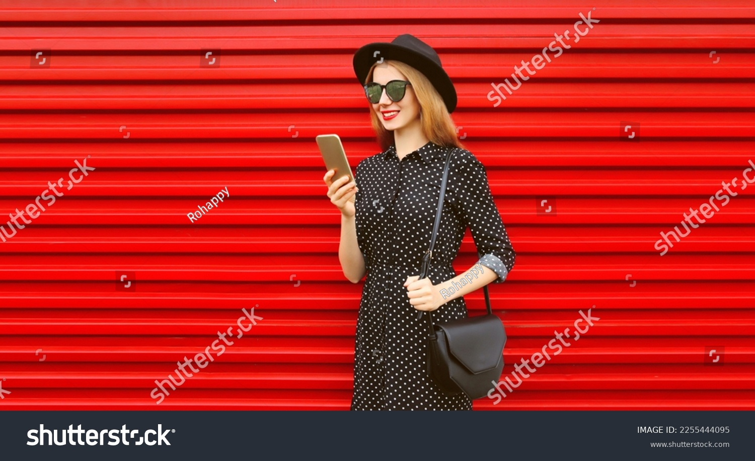 Portrait of stylish smiling woman with smartphone wearing black round hat on red background #2255444095