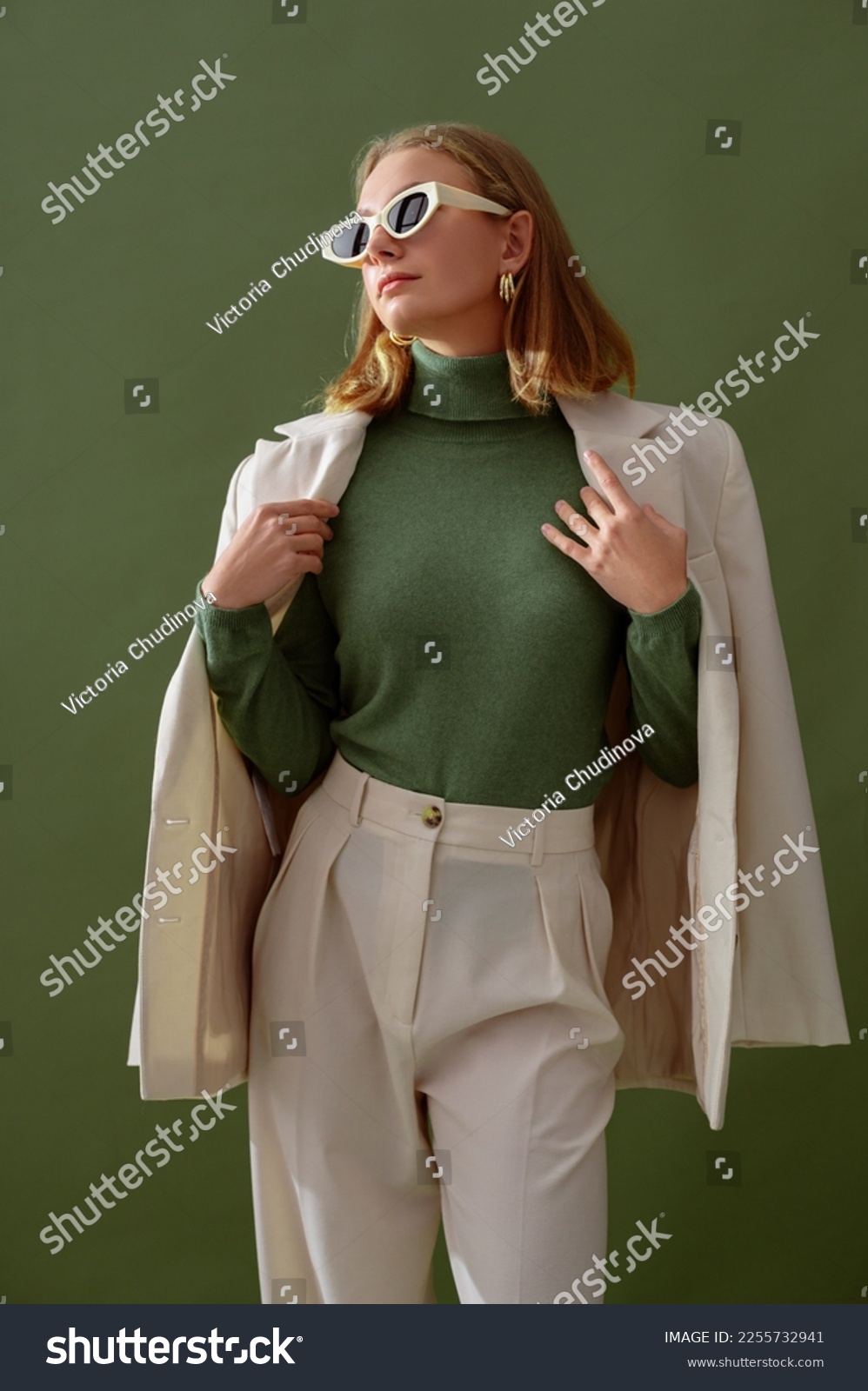 Fashionable confident woman wearing elegant white suit with blazer, trousers, cashmere turtleneck sweater, trendy cat eye sunglasses, posing on green background. Studio fashion portrait #2255732941