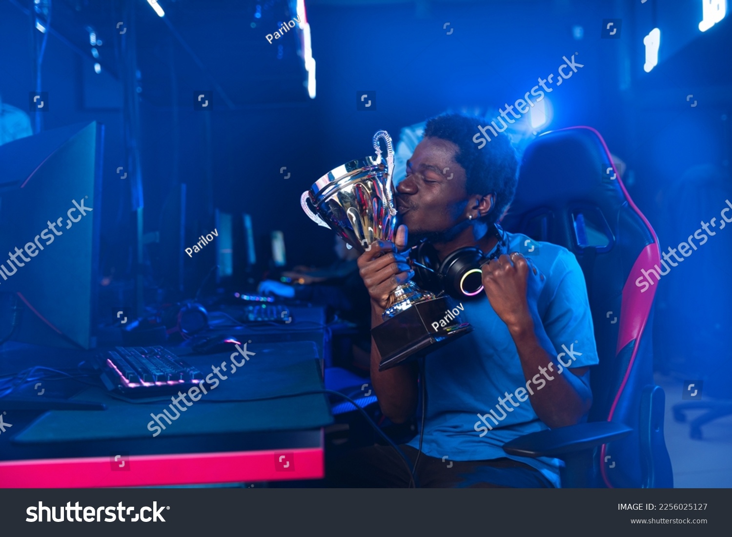 Happy American African Professional gamer, winner rejoices in victory of online games tournaments pc computer with headphones, Blurred red and blue background. #2256025127