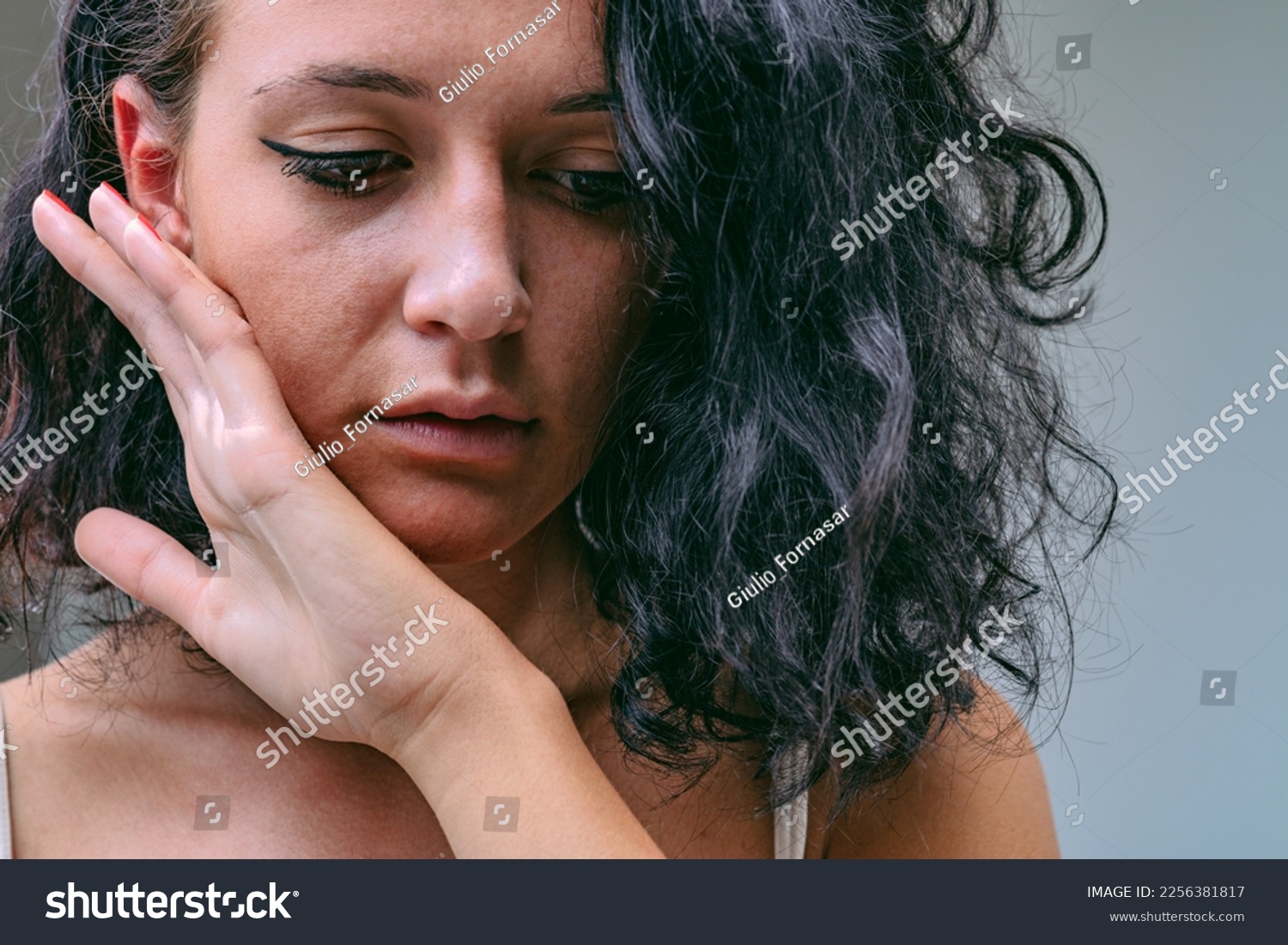 Frontal close-up portrait of young woman bringing her hand to touch the skin of her cheek and chin with the back. She may be feeling how smooth it is, but she may be sad and afraid of a slap. #2256381817