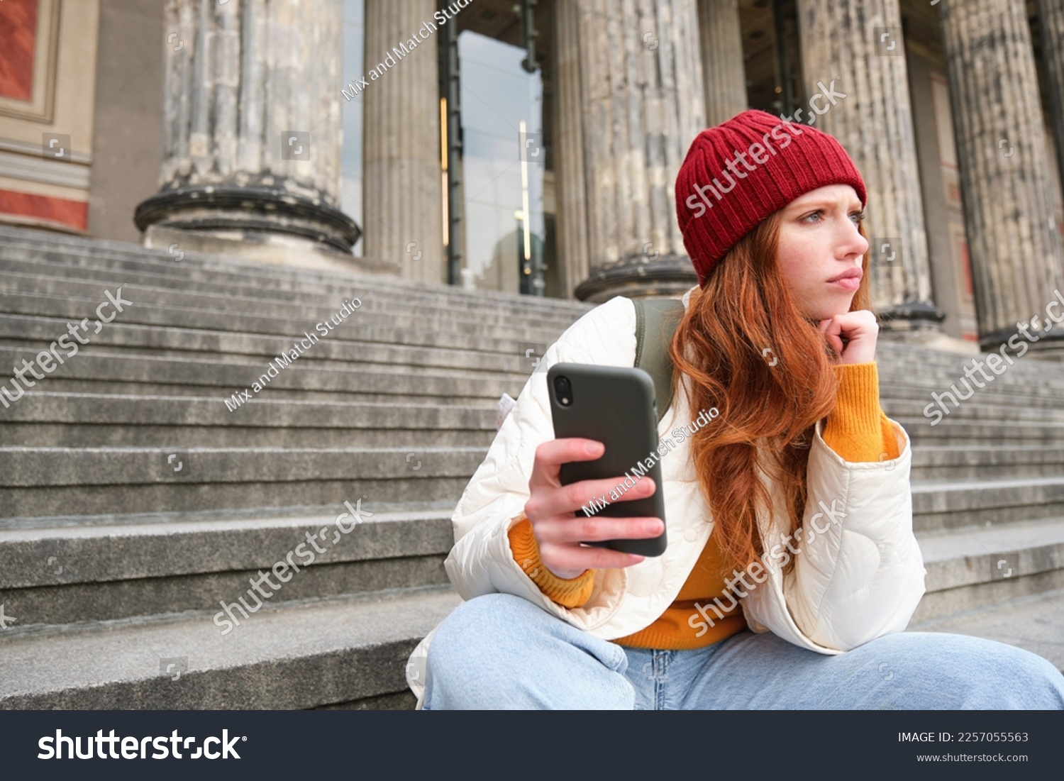 Portrait of young redhead woman with complicated face, sits on street stairs in red hat, holds smartphone and frowns thoughtful, feels uneasy. #2257055563