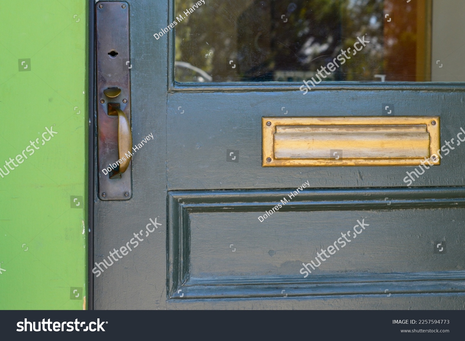 A dark green wooden door with half glass pane window, and a wooden panel. The vintage door has a brass door handle, a keyhole, and a letter plate. The trim of the building is vibrant green color.  #2257594773