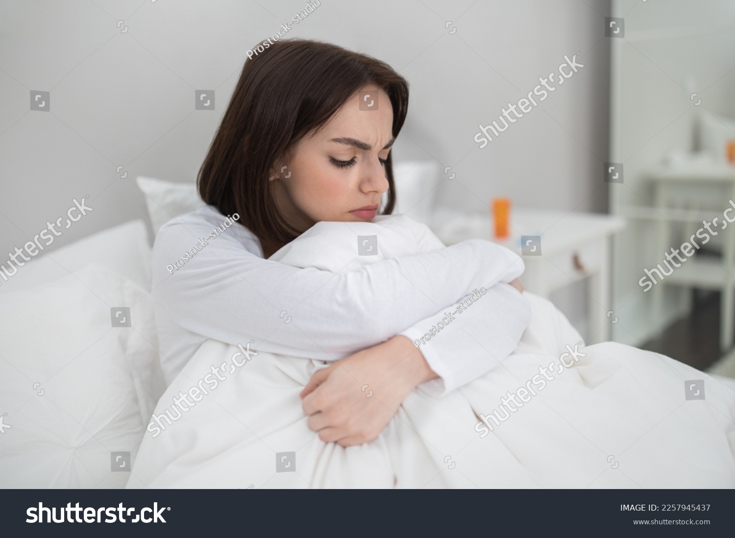 Young sick unhappy brunette woman sitting in bed at home, lady got cold, flu or coronavirus, suffering from period cramps or headache, medicine on bedside table, copy space #2257945437