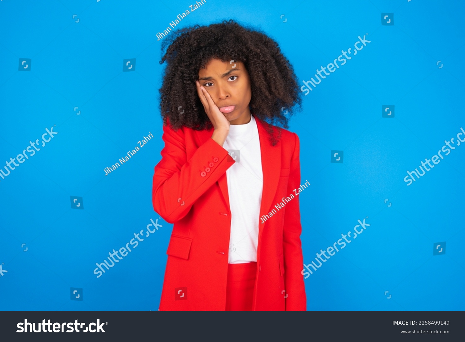 Sad lonely young businesswoman with afro hairstyle wearing red over blue background touches cheek with hand bites lower lip and gazes with displeasure. Bad emotions #2258499149