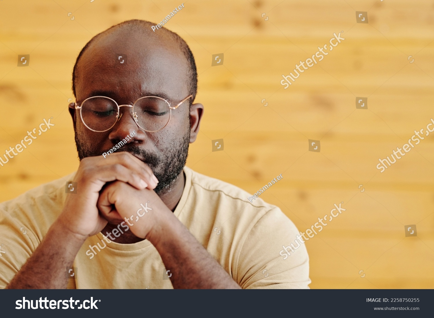 Young black man in eyeglasses keeping eyes closed and clenched hands by his mouth while concentrating on solving serious problem #2258750255