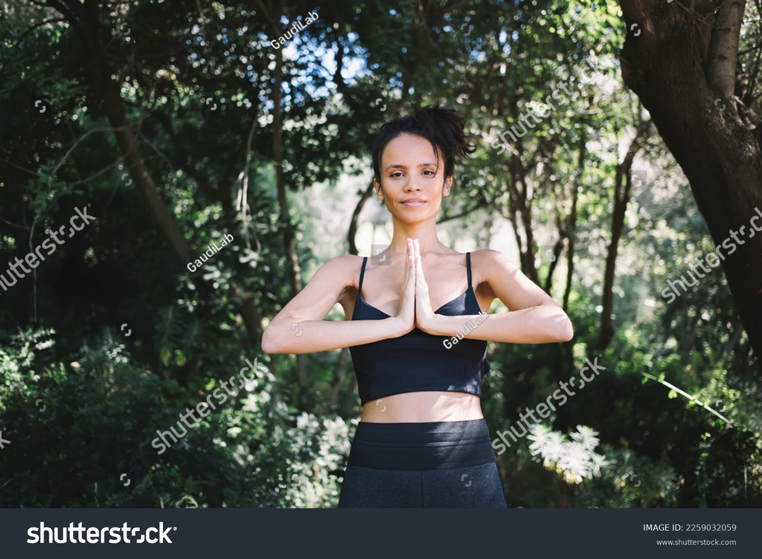 Peaceful young ethnic female in activewear standing with hands clasped and practicing namaste gesture while looking at camera in forest #2259032059
