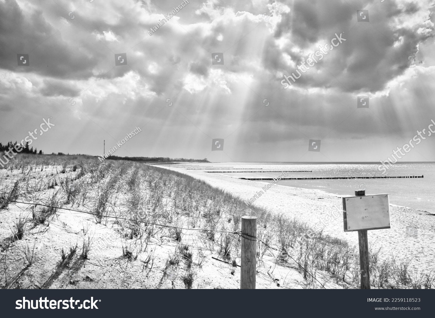 Sun rays shining through dense clouds on the beach of the Baltic Sea taken in black and white. Sign at the beach crossing. Groynes on the coast. Sandy beach in Zingst. Landscape photo from Germany #2259118523
