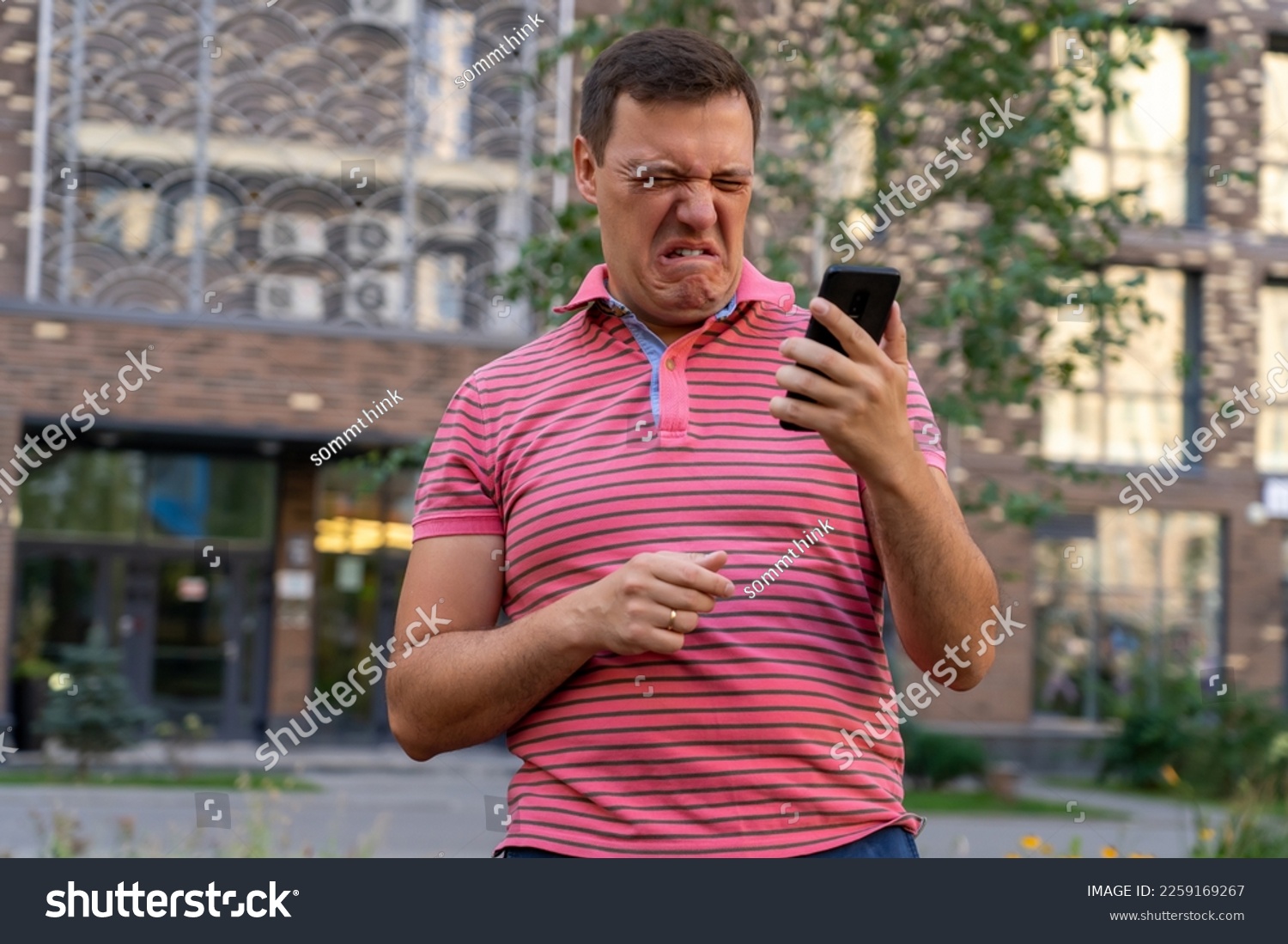 Dissatisfied millennial man in pink T-shirt, chatting on phone, looking at gadget with offended and disgusted expression, receiving bad message. #2259169267