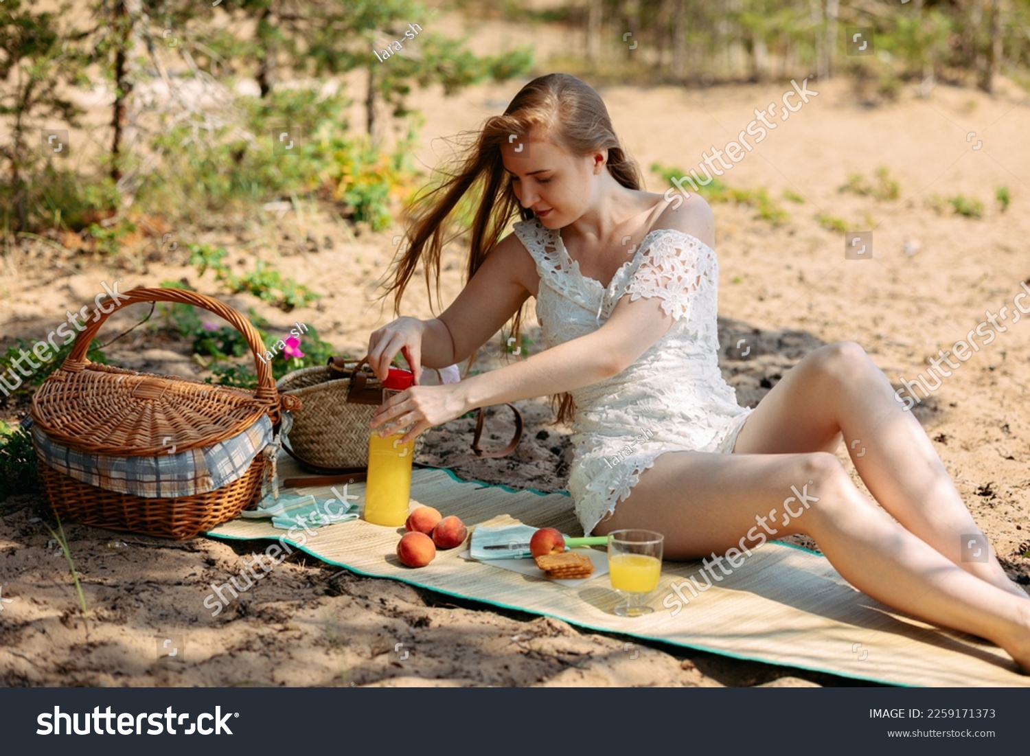 Romantic blonde woman in straw hat having picnic on sea beach. Fresh fruits, juice, cookies and peaches. High quality photo #2259171373