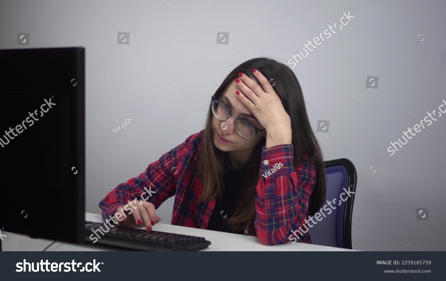 An IT specialist sits wearily in the office in front of a computer. A young girl lazily types on the keyboard. Girl with glasses and a red shirt. #2259185759