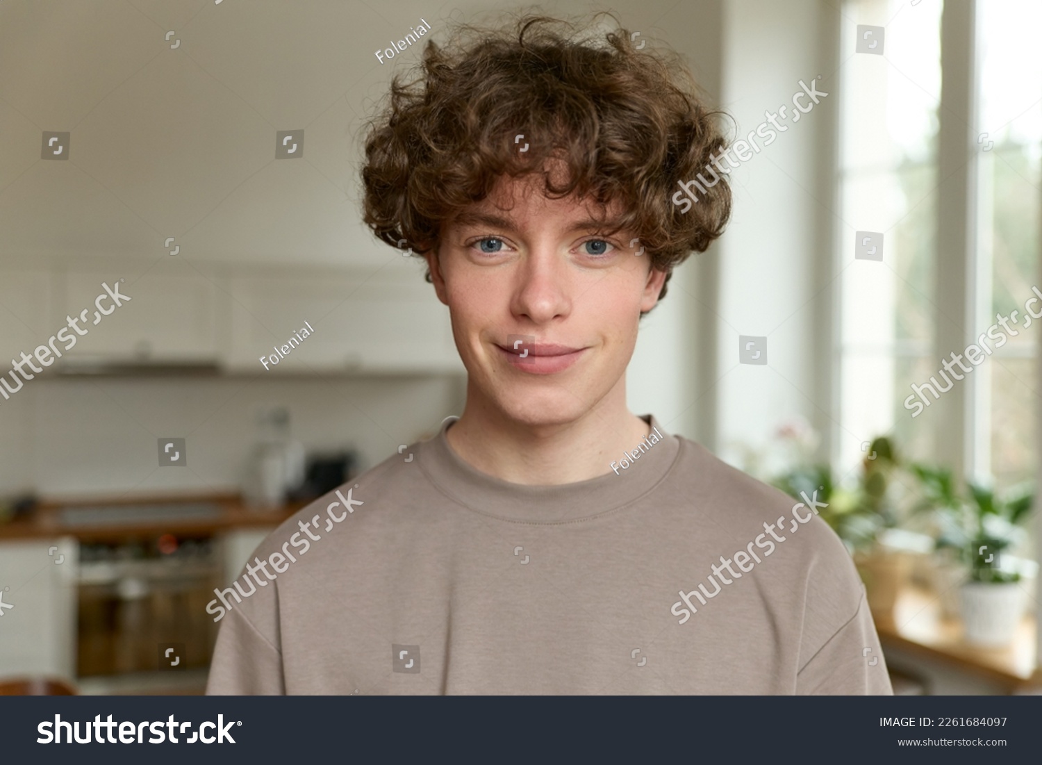 Close up of a young handsome positive curly man with blue eyes with a smiling face wearing casual beige t-shirt looking at camera and standing alone in morning in the modern kitchen at home. #2261684097
