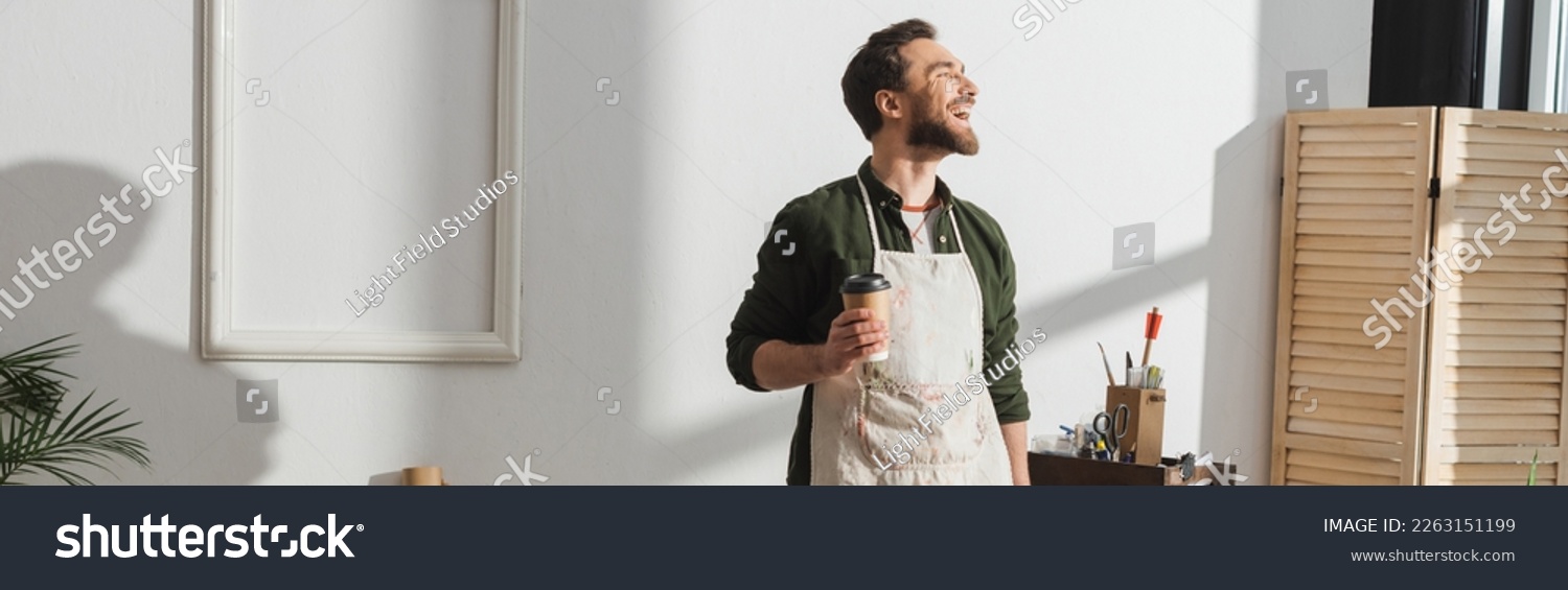 Smiling bearded repairman in apron holding paper cup in workshop, banner #2263151199