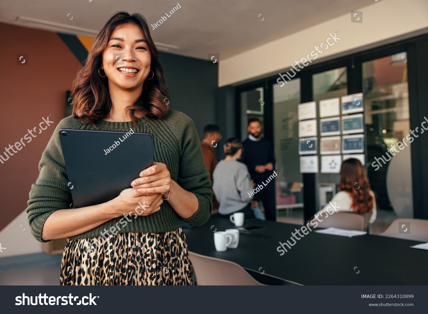 Cheerful young businesswoman smiling at the camera while holding a digital tablet. Happy young businesswoman standing in a boardroom with her colleagues in the background. #2264310899