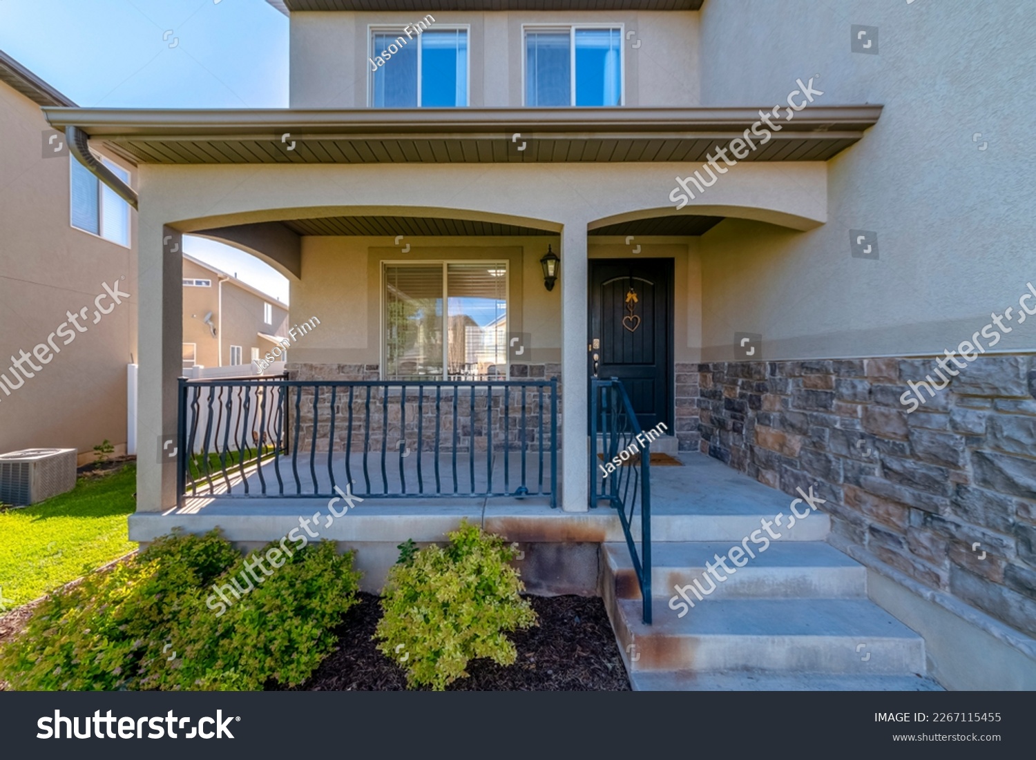House entrance at the side of a wall with stucco and stone veneer. House exterior with railings on the porch at front of the window beside the black front door with hanging ornaments. #2267115455
