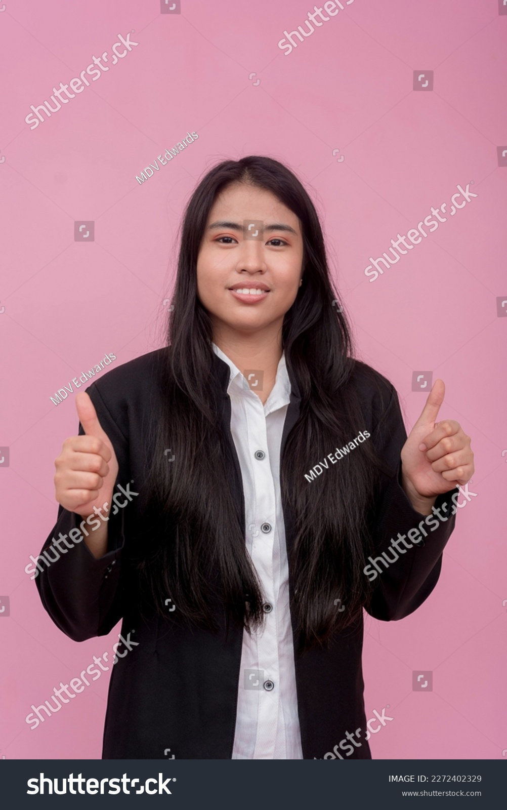 A young southeast asian female employee in a black jacket with arms crossed. Isolated on a light pink background. #2272402329