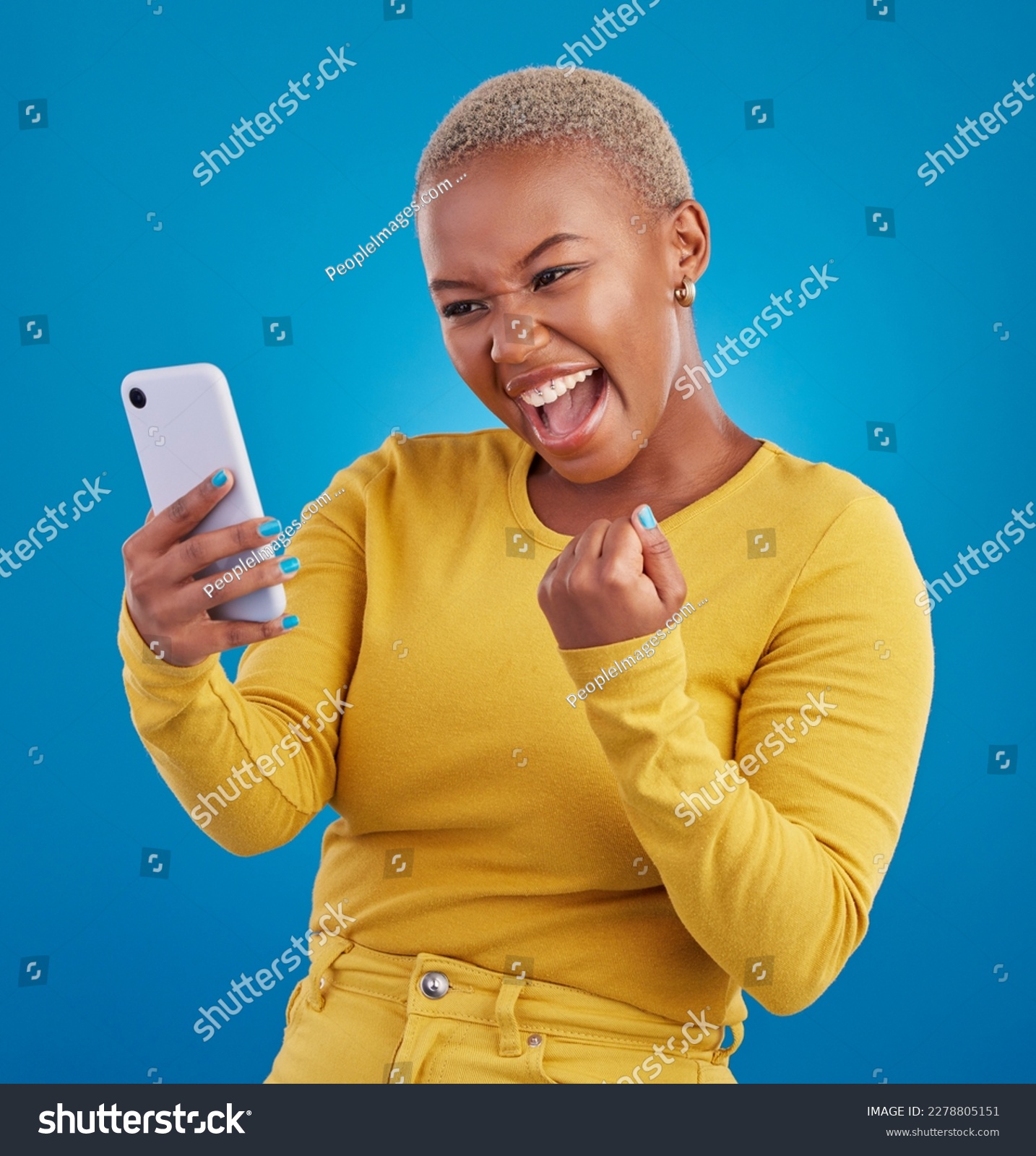 Black woman, phone and fist celebration in studio, blue background and winning online prize. Happy female model celebrate mobile promotion, bonus and excited for deal, success and competition winner #2278805151