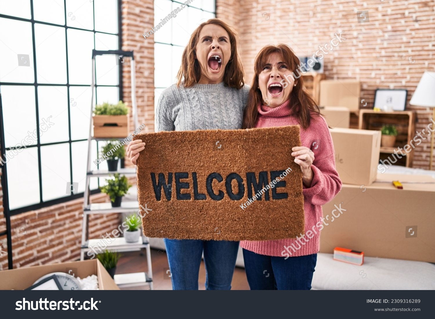 Mother and daughter holding welcome doormat angry and mad screaming frustrated and furious, shouting with anger looking up.  #2309316289