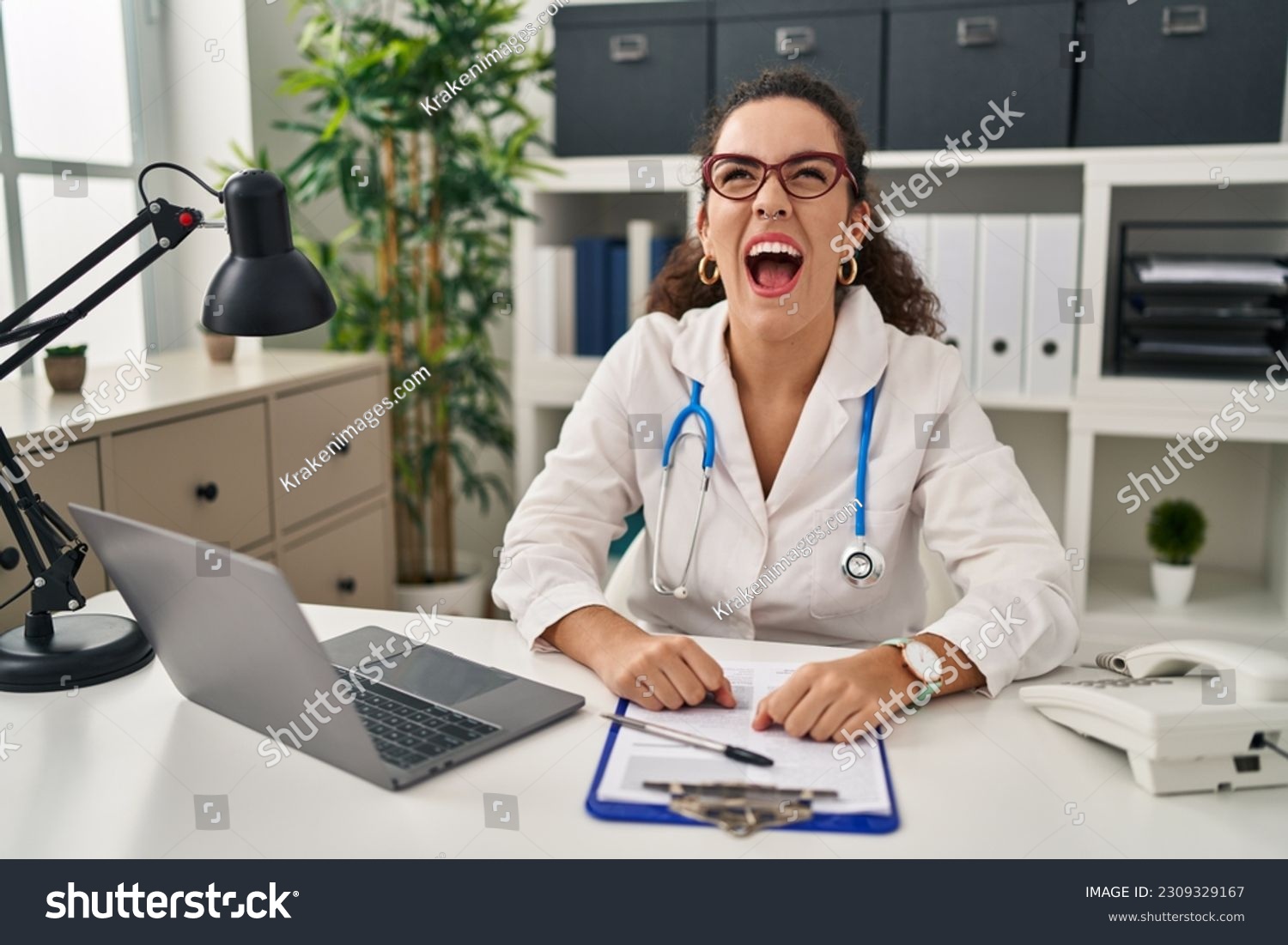 Young hispanic woman wearing doctor uniform and stethoscope angry and mad screaming frustrated and furious, shouting with anger. rage and aggressive concept.  #2309329167