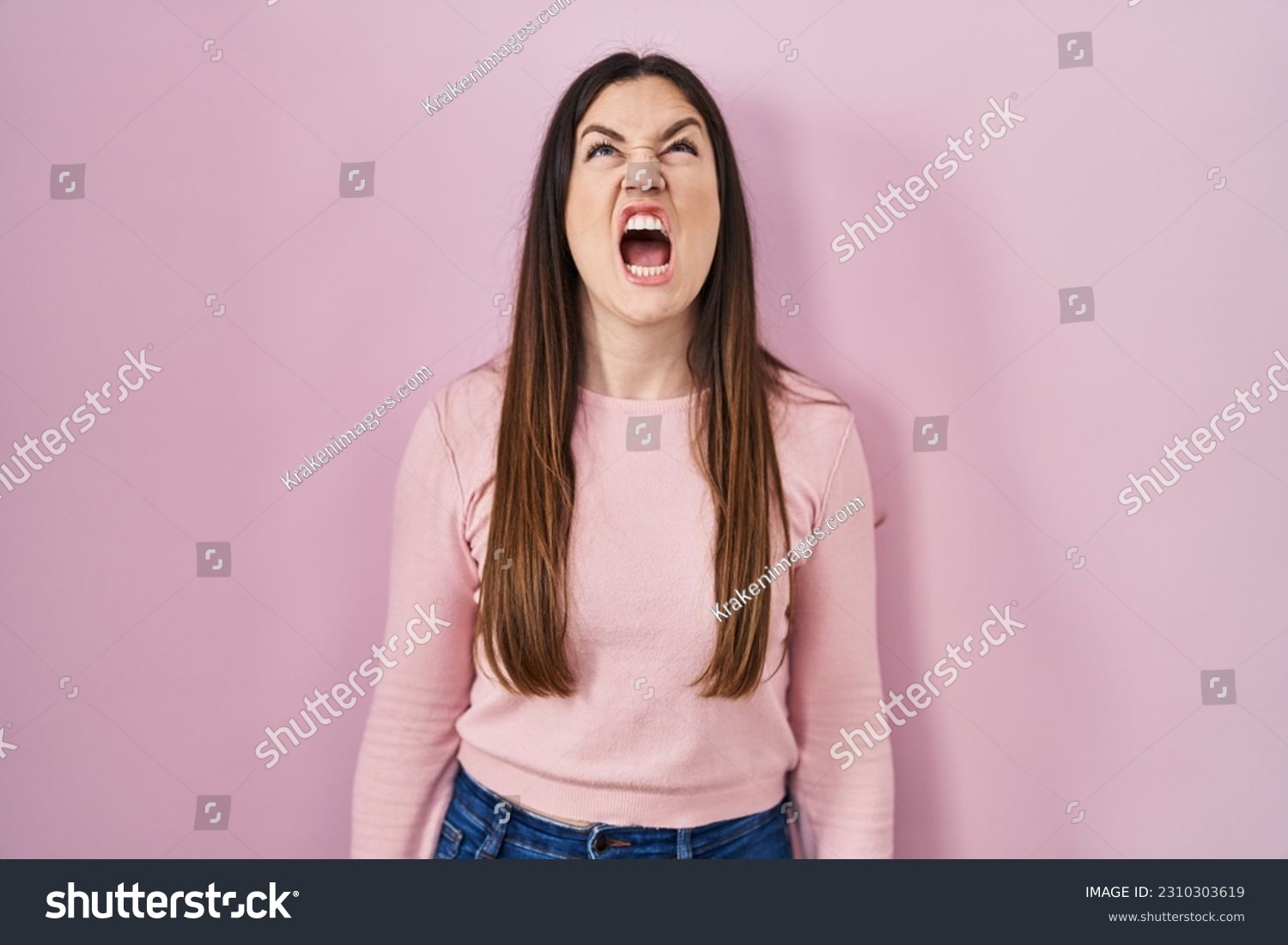 Young brunette woman standing over pink background angry and mad screaming frustrated and furious, shouting with anger. rage and aggressive concept.  #2310303619